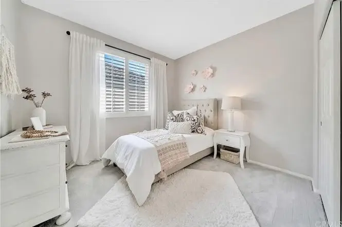 Bedroom with single bed, white and beige quilt, bedside table with lamp, window with curtains, and rug.