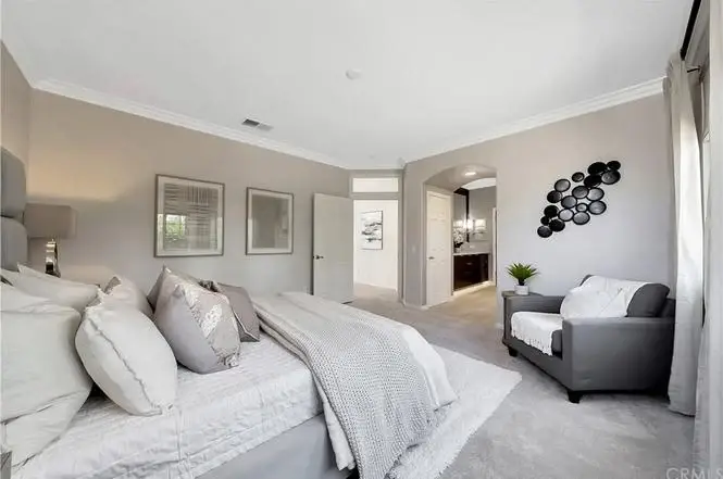 Bedroom with bed, light bedding, gray chair, wall art, soft lights, and doorway to bathroom.