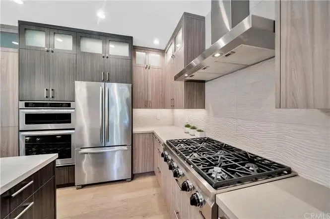 Kitchen with stainless steel fridge, oven, gas range, wood cabinets, and textured backsplash.