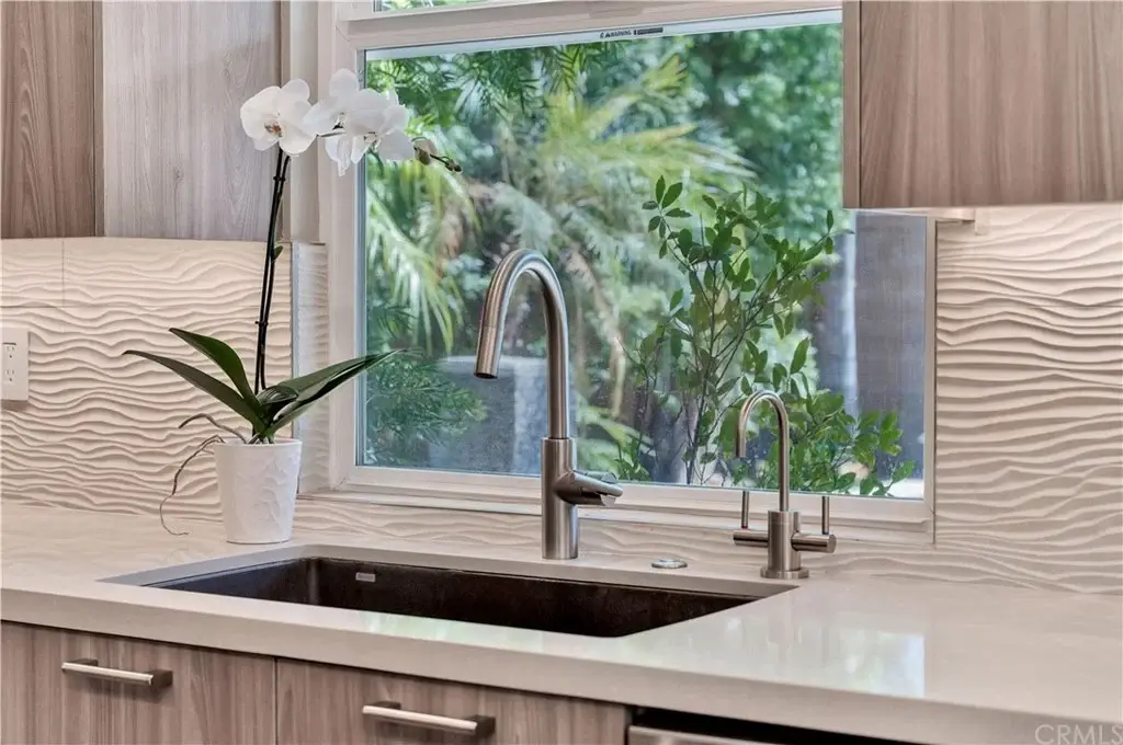 Kitchen sink with sleek faucet, window view of plants, white orchid, and textured backsplash.