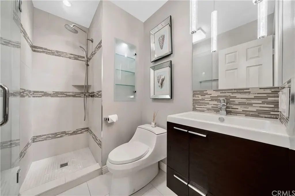 Bathroom with walk in glass shower, toilet, vanity sink, gray walls, wall art, and lights.