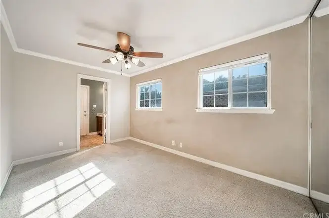 Empty bedroom with beige walls, two windows, ceiling fan, and soft carpet floor.