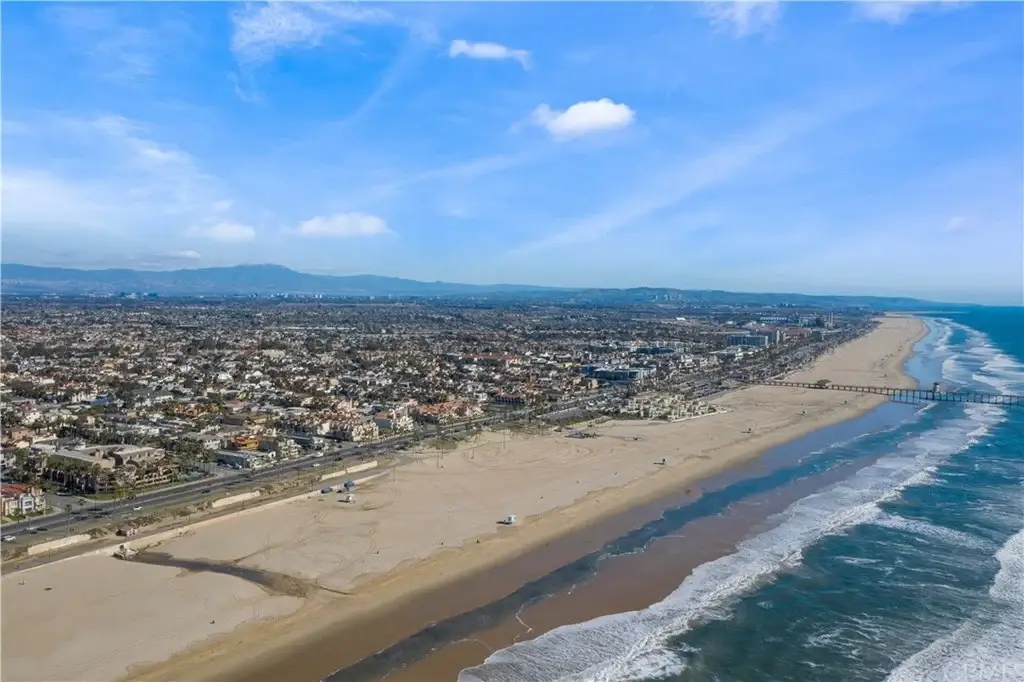 Aerial view of beach shoreline with waves, pier in the distance, and city along the coast.