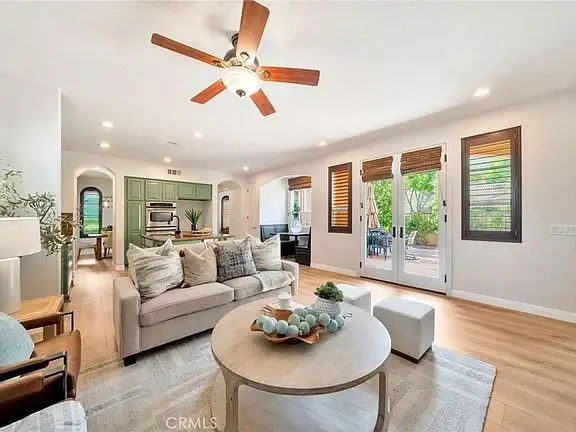 Living room with ceiling fan, light-colored seating, round coffee table, large windows, and greenery outside