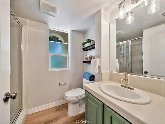 Bathroom with toilet, sink, mirror, light-colored walls, window, green cabinets, and shelves with decorative items