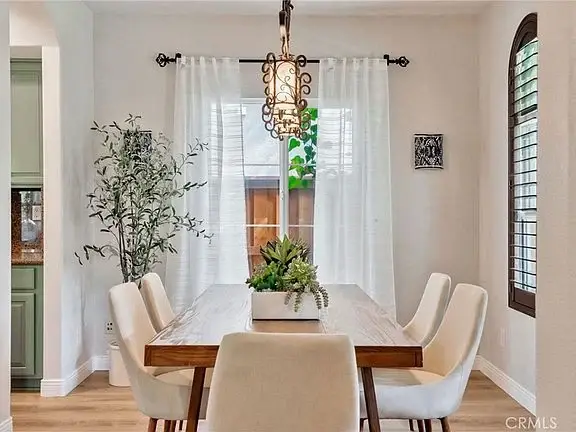 Dining area with wooden table, white chairs, potted plants, and window with curtains