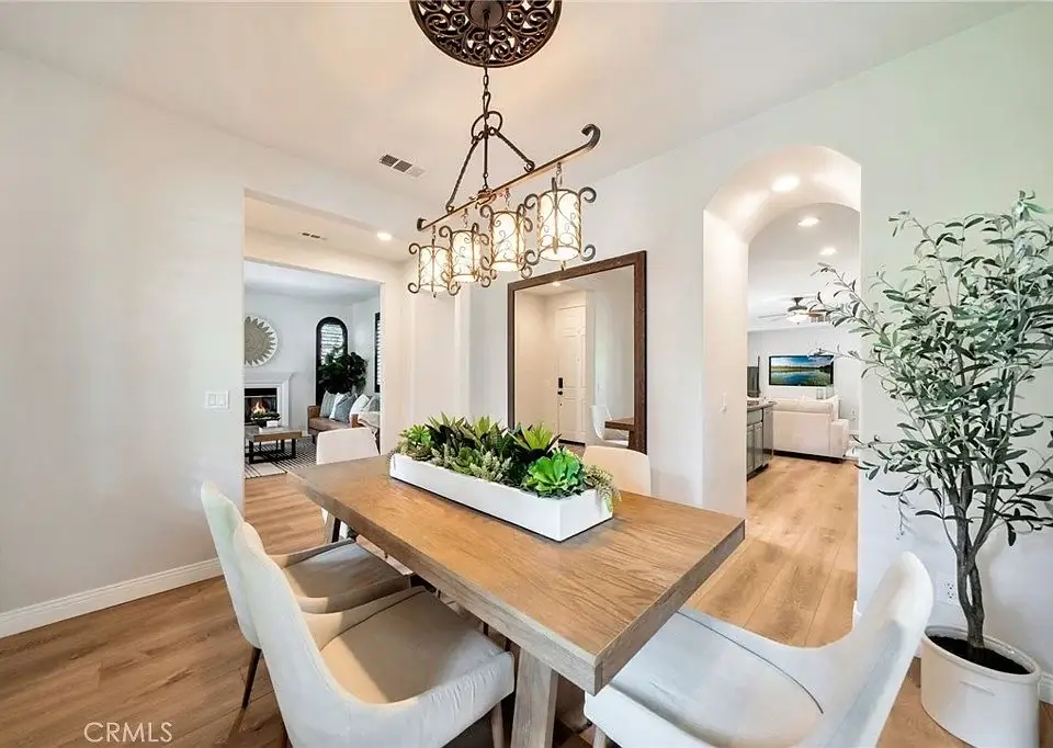 Dining room with wooden table, white chairs, chandelier, plants, and open view to room with fireplace