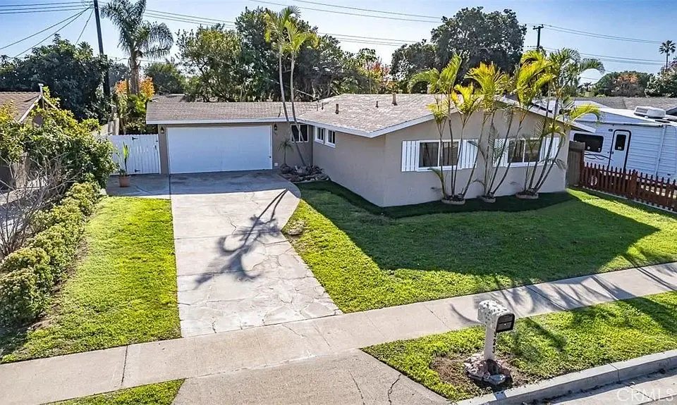 Single-story California real estate house with a driveway, garage, green grass, and palm trees, with other houses in the background.
