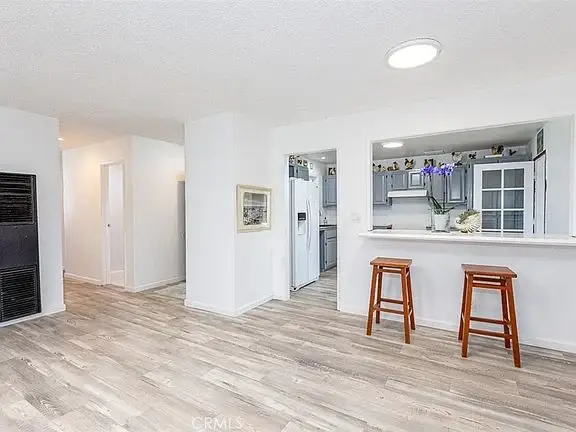 Open living space with kitchen bar, two wooden stools, white walls, light floor, and appliances.