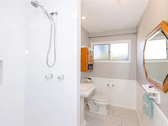Bathroom with light gray walls, shower, toilet, sink, small window, and wooden mirror.