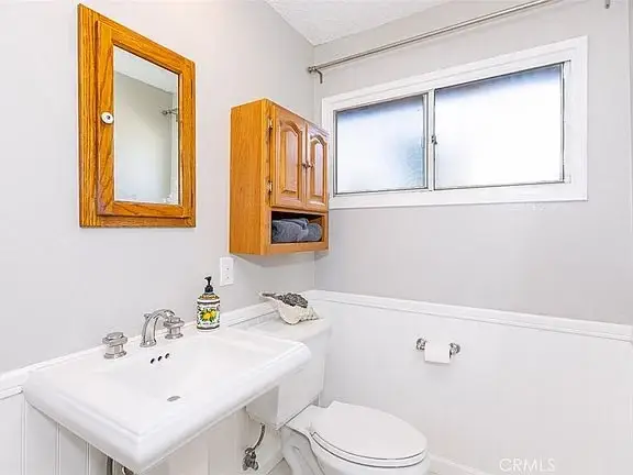 bathroom with white sink, wooden cabinet, toilet, mirror, and window.