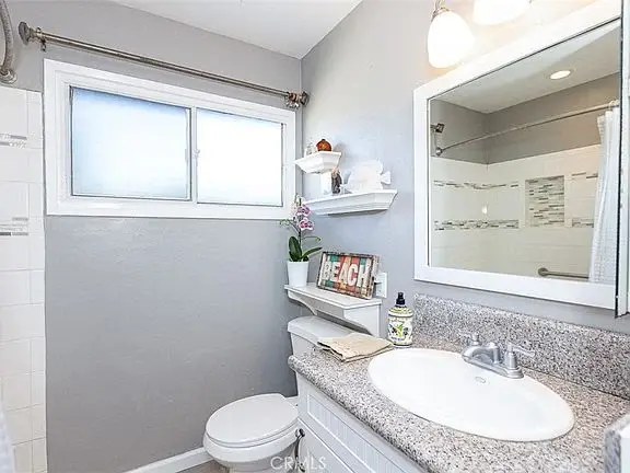 Bathroom with light gray walls, white vanity with sink, toilet, window, and shower with glass doors.