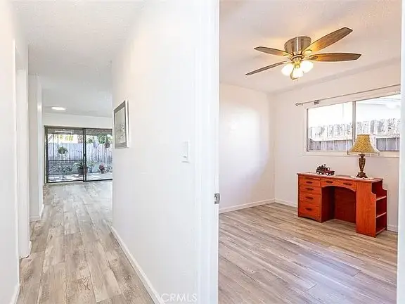Hallway with light wood floor leading to a room with a wooden desk, ceiling fan, window, and view of an outdoor area.