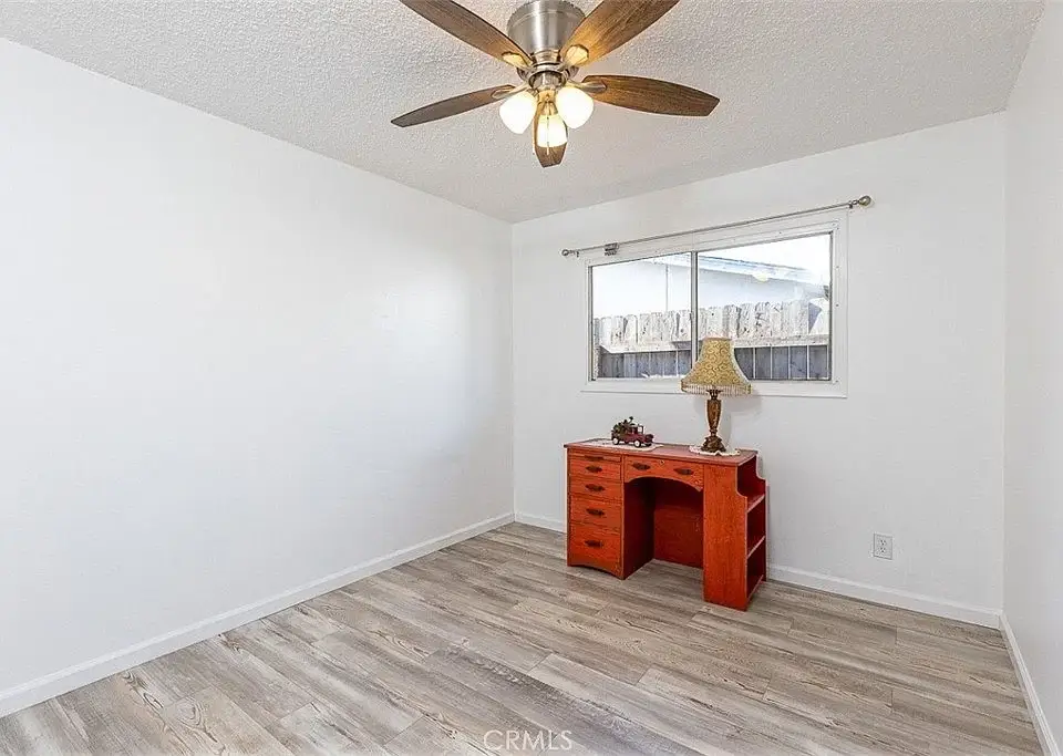 Bright room with white walls, wood laminate floor, ceiling fan, and small wooden desk under a window.