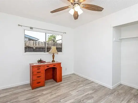 Empty room with white walls, wood‑like floor, ceiling fan with lights, window, and a small desk in the corner.