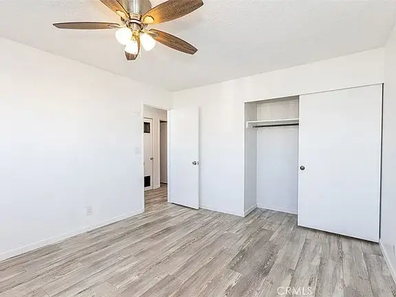 Empty room with light walls, light wood floor, ceiling fan, closet with sliding doors, and open doorway.