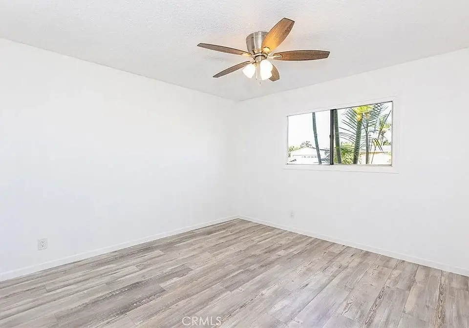 Empty room with white walls, light wooden floor, ceiling fan, and window with palm trees outside.