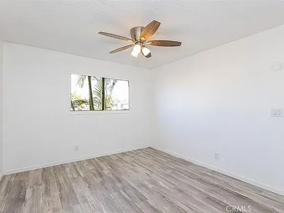 empty room with white walls, light wooden floor, ceiling fan, and window showing palm trees outside.