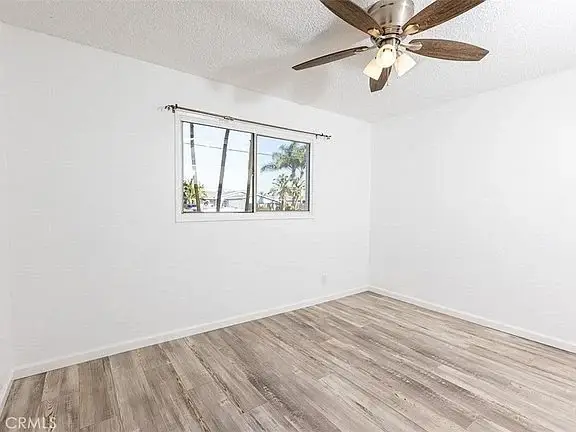 empty room with white walls, wooden floor, ceiling fan, and window showing trees outside.