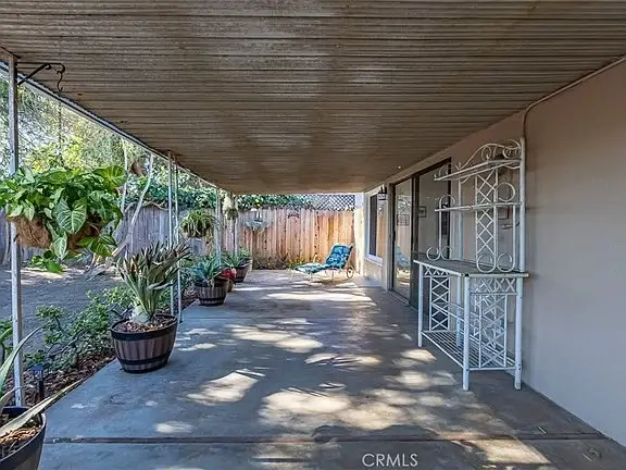 Covered outdoor patio with potted plants, a blue chair, greenery, and a wooden fence.