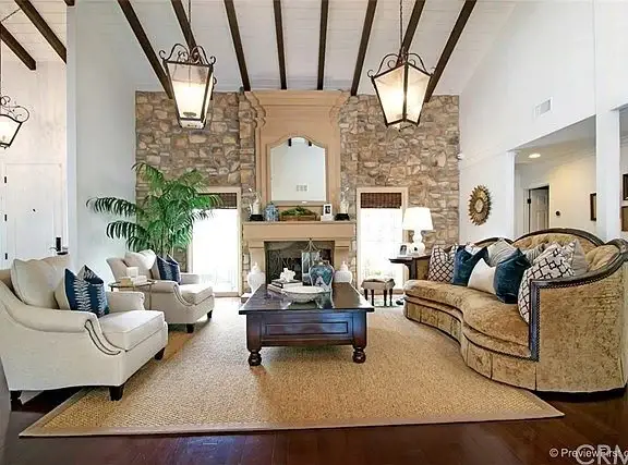 Living room with stone fireplace, wood beam ceiling, seating with patterned cushions, and rug.