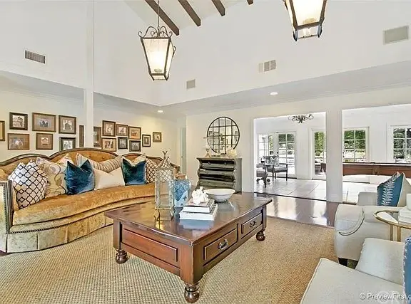 Living room with stone wall, fireplace with mirror, cream and patterned sofas, coffee table, and windows.