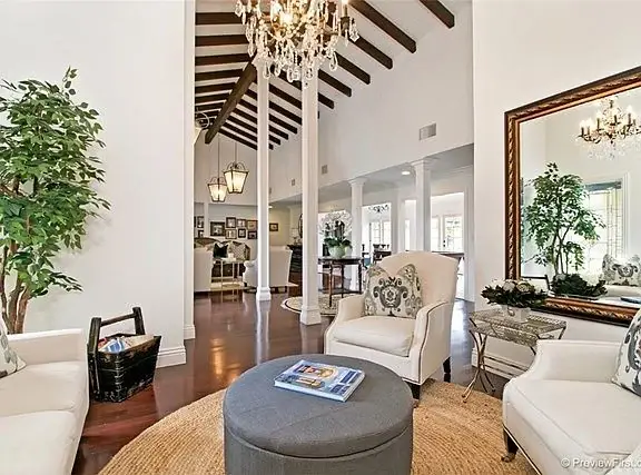 Living room with high ceiling, wood beams, white chairs, round ottoman, chandelier, plants, and large windows.