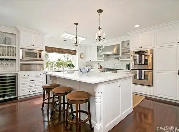 Kitchen with white cabinets, stainless steel appliances, large island with three wooden stools, and hardwood floor.
