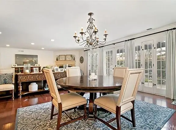 Dining room with round wooden table, four chairs, chandelier, large windows, hardwood floor, and rug.
