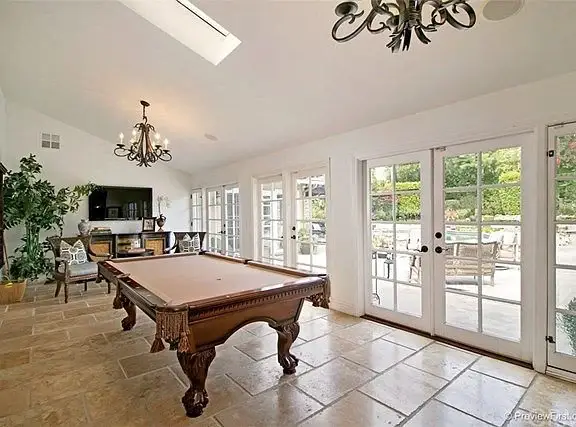 Room with pool table, chandelier, chairs, TV, glass doors, stone tile floor, and plants.