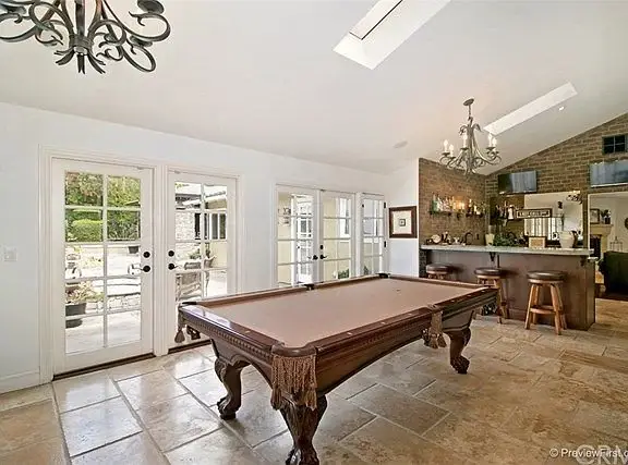 Room with billiard table, bar area, chandeliers, stone walls, and large windows and doors.
