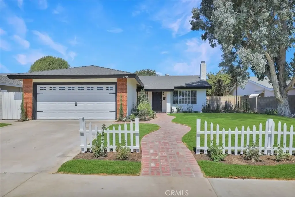 House California real estate with brick path, white fence, two car garage, green lawn, trees, and blue sky.